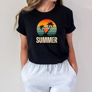 Peut inclure: Un t-shirt noir avec un motif r&eacute;tro de palmiers et un coucher de soleil. Le motif est dans des tons d'orange, jaune et bleu. Le texte "SUMMER" est imprim&eacute; sous le motif.