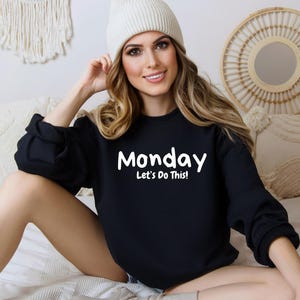 May include: Black sweatshirt with white text that reads "Monday Let's Do This!" A woman is wearing the sweatshirt and a white beanie.