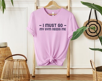 May include: A light pink t-shirt with the text "I must go - my gym needs me" printed in dark blue.