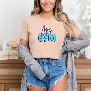 May include: A beige t-shirt with the text "Mrs. GYM" in blue bubble letters. The model is wearing denim shorts and a gray cardigan. The shirt is a crew neck style.