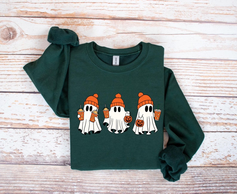 K&ouml;nnte beinhalten: Ein dunkelgr&uuml;ner Pullover mit einem Motiv von drei Geistern, die orangefarbene H&uuml;te tragen und K&uuml;rbisf&ouml;rmige Eimer und orangefarbene Getr&auml;nke halten.