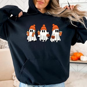 May include: A black hooded sweatshirt featuring a graphic of three white ghosts wearing orange knit beanies. Each ghost holds an orange beverage cup, and one ghost also carries a small orange jack-o'-lantern pumpkin bucket.