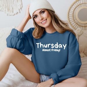 May include: A woman wearing a blue sweatshirt with the text "Thursday Almost Friday!" in white. She is sitting on a bed with a white beanie on her head.