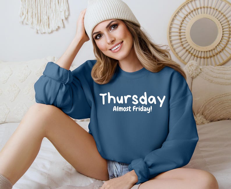 May include: A woman wearing a blue sweatshirt with the text "Thursday Almost Friday!" in white. She is sitting on a bed with a white beanie on her head.
