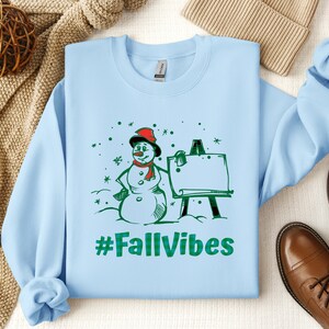 May include: A light blue crewneck sweatshirt featuring a graphic of a snowman wearing a red scarf and hat standing next to an easel. The design includes green snowflakes and the text #FallVibes in a green plaid pattern printed on the front.