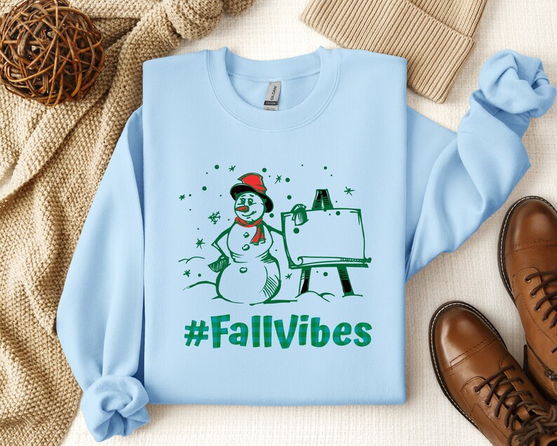 May include: A light blue crewneck sweatshirt featuring a graphic of a snowman wearing a red scarf and hat standing next to an easel. The design includes green snowflakes and the text #FallVibes in a green plaid pattern printed on the front.