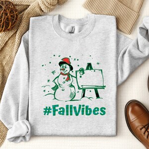 May include: A light gray crewneck sweatshirt featuring a graphic of a snowman wearing a red hat and scarf, standing next to an easel. The design includes green snowflakes and the text #FallVibes in a green plaid pattern printed on the front.