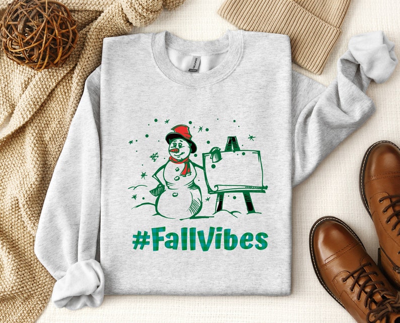 May include: A light gray crewneck sweatshirt featuring a graphic of a snowman wearing a red hat and scarf, standing next to an easel. The design includes green snowflakes and the text #FallVibes in a green plaid pattern printed on the front.