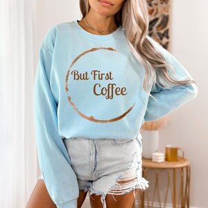 May include: A light blue sweatshirt with the text "But First Coffee" in brown. The design includes a coffee ring stain around the text. The sweatshirt is a casual style.