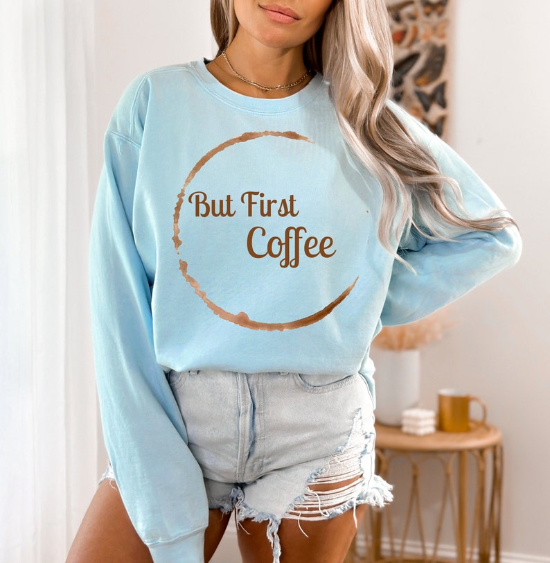 May include: A light blue sweatshirt with the text "But First Coffee" in brown. The design includes a coffee ring stain around the text. The sweatshirt is a casual style.