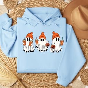 May include: A light blue hooded sweatshirt featuring a graphic of three white ghosts wearing orange knit beanies. The ghosts are holding orange drinks and small orange pumpkin-shaped buckets for collecting treats.