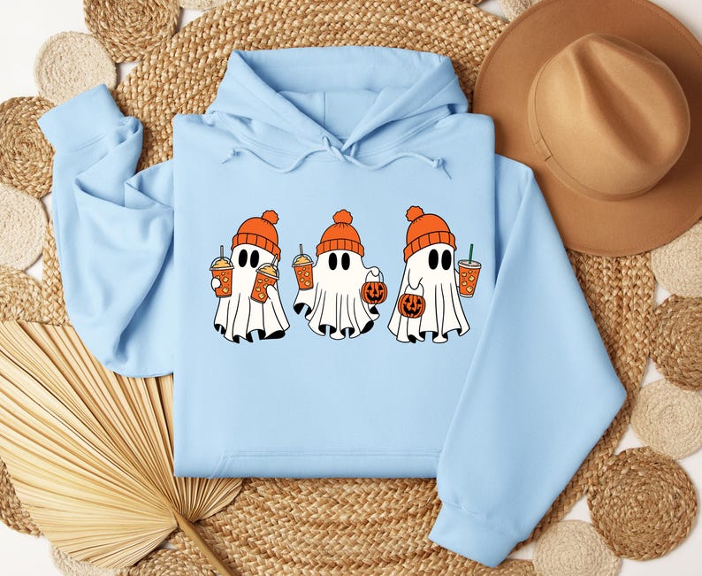 May include: A light blue hooded sweatshirt featuring a graphic of three white ghosts wearing orange knit beanies. The ghosts are holding orange drinks and small orange pumpkin-shaped buckets for collecting treats.