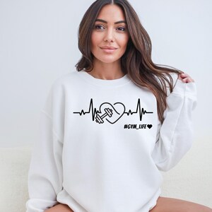 Peut inclure: Sweat-shirt blanc avec un graphique noir d'un cœur avec une haltère à l'intérieur, une ligne de battement de cœur et le texte "#GYM_LIFE".