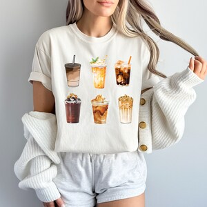 May include: A white t-shirt with a graphic of six different iced coffee drinks. The drinks are all in clear cups with different toppings and flavors.