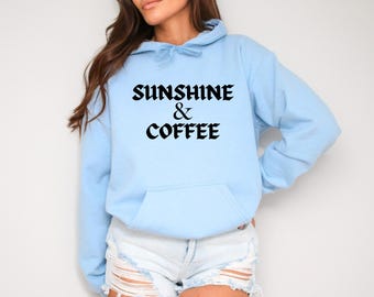 May include: A light blue hooded sweatshirt featuring a front kangaroo pocket and the black text "SUNSHINE & COFFEE" printed in a bold, gothic-style font across the chest area. The garment includes a matching drawstring hood.