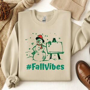 May include: A beige crewneck sweatshirt featuring a graphic print of a snowman wearing a red hat and scarf, standing next to an easel. The design includes green snowflakes and the text #FallVibes in a green and blue plaid pattern font.