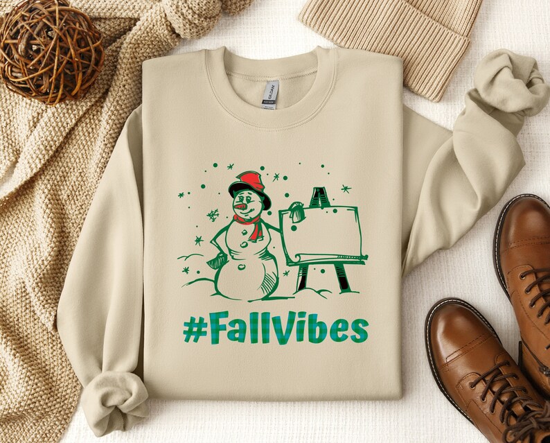 May include: A beige crewneck sweatshirt featuring a graphic print of a snowman wearing a red hat and scarf, standing next to an easel. The design includes green snowflakes and the text #FallVibes in a green and blue plaid pattern font.