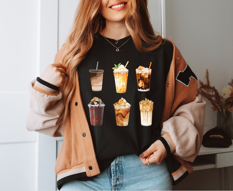May include: A black t-shirt with a graphic of six different iced coffee drinks. The drinks are all in clear cups with different toppings and flavors. The shirt is being worn by a person with long brown hair.