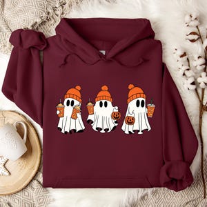 May include: A deep maroon hooded sweatshirt featuring a graphic print of three white ghosts wearing orange knit beanies. The ghosts are holding orange pumpkin-shaped buckets and orange cups with straws, creating a festive autumn-themed design.