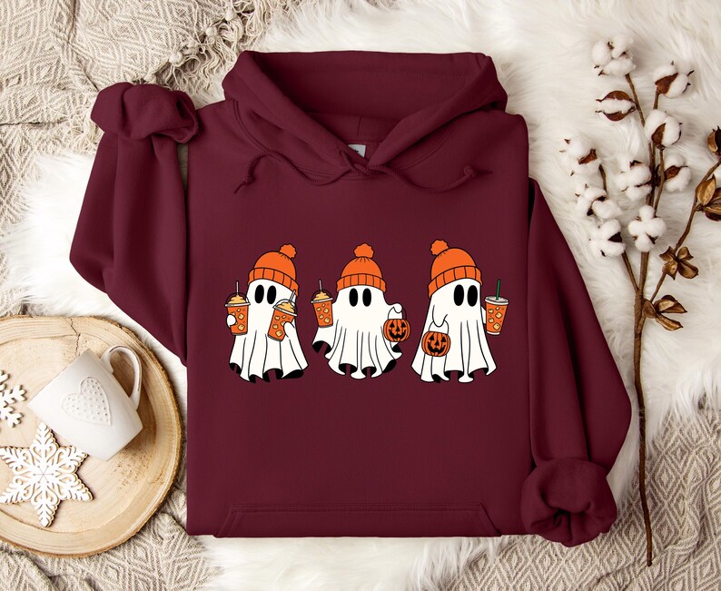 May include: A deep maroon hooded sweatshirt featuring a graphic print of three white ghosts wearing orange knit beanies. The ghosts are holding orange pumpkin-shaped buckets and orange cups with straws, creating a festive autumn-themed design.