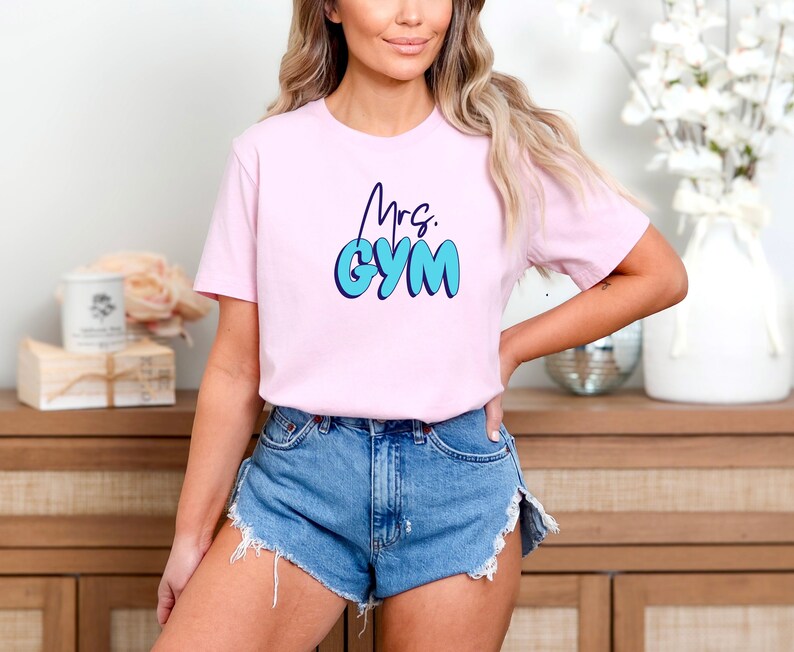 May include: A light pink t-shirt with the text "Mrs. GYM" in a blue bubble font. The shirt is short-sleeved and appears to be made of a soft material. The model is wearing denim shorts.