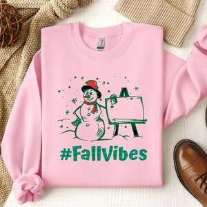 May include: A light pink long-sleeve sweatshirt featuring a graphic print of a snowman wearing a red hat and scarf standing next to an easel. The text #FallVibes is printed in green below the snowman. The design includes green and red accents.