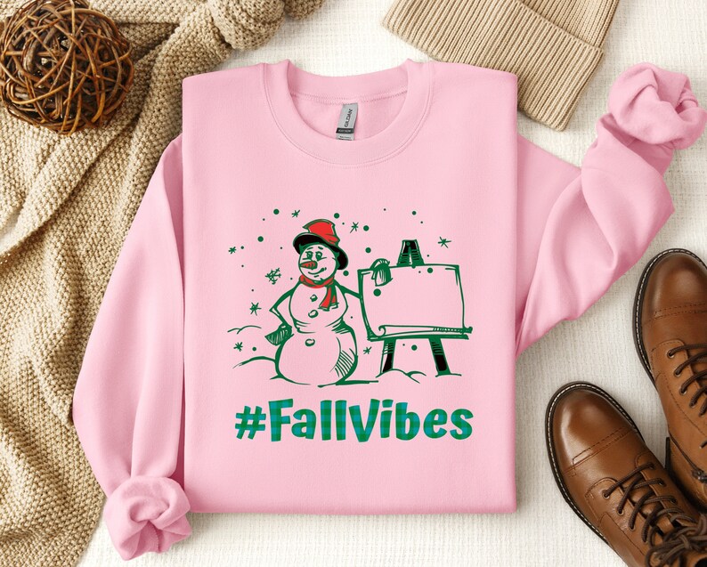 May include: A light pink long-sleeve sweatshirt featuring a graphic print of a snowman wearing a red hat and scarf standing next to an easel. The text #FallVibes is printed in green below the snowman. The design includes green and red accents.