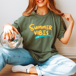 May include: A woman wearing a green t-shirt with the text "Summer Vibes only" in yellow and blue lettering. She is holding a silver disco ball.