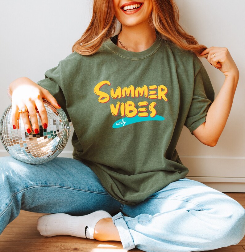 May include: A woman wearing a green t-shirt with the text "Summer Vibes only" in yellow and blue lettering. She is holding a silver disco ball.