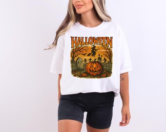 May include: White t-shirt with a Halloween graphic. The design features an orange pumpkin with a carved face, a witch flying on a broom, and a graveyard scene with the word "Halloween" at the top. The shirt is a casual, oversized fit.