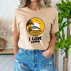 May include: A beige t-shirt featuring a graphic of a palm tree, sun, and birds within a yellow circle. The text "i love summer" is printed beneath the circle.