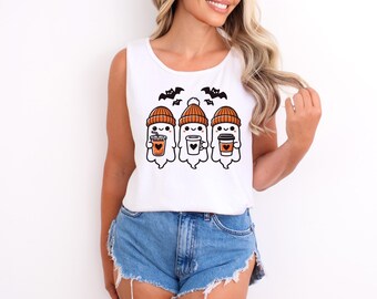 May include: A white sleeveless tank top featuring a graphic print of three cartoon ghosts wearing orange beanies. Each ghost holds a different beverage, including a cup with a heart, and two small black bats are flying above them.