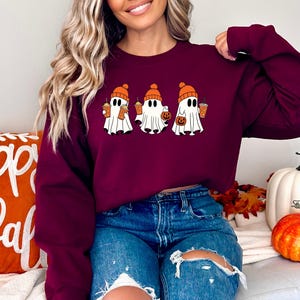 K&ouml;nnte beinhalten: Ein maroonfarbener Pullover mit einem Aufdruck von drei Geistern, die orangefarbene H&uuml;te tragen und orangefarbene Becher und einen K&uuml;rbis halten. Die Geister tragen orangefarbene H&uuml;te und halten orangefarbene Becher und einen K&uuml;rbis. Der Pullover ist im Cropped-Stil gehalten und wird von einer Person mit langen braunen Haaren getragen.