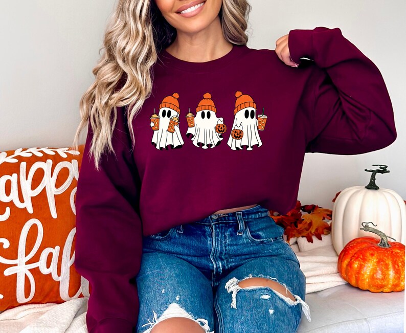 K&ouml;nnte beinhalten: Ein maroonfarbener Pullover mit einem Aufdruck von drei Geistern, die orangefarbene H&uuml;te tragen und orangefarbene Becher und einen K&uuml;rbis halten. Die Geister tragen orangefarbene H&uuml;te und halten orangefarbene Becher und einen K&uuml;rbis. Der Pullover ist im Cropped-Stil gehalten und wird von einer Person mit langen braunen Haaren getragen.