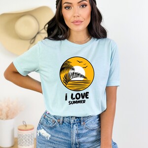 May include: A light blue t-shirt with a yellow circle graphic of a palm tree, sun, and ocean waves. The text "i love summer" is printed below the graphic.