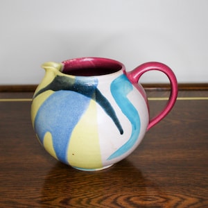 May include: A round ceramic jug with a pink handle and abstract blue, yellow and black painted designs.