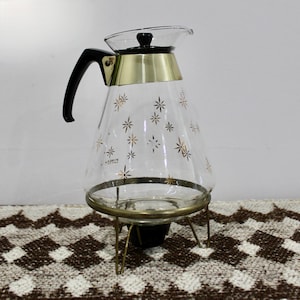 May include: Vintage glass coffee carafe with a black handle and lid, and a gold-toned metal band. The clear glass is decorated with gold starburst designs. The carafe sits on a gold-toned metal stand with three legs.
