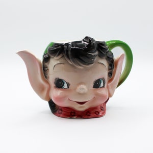 May include: A novelty ceramic teapot in the shape of an elf's face. The teapot has black hair, large blue eyes, pink cheeks and ears, a red collar, and a green handle. The spout is on the left side.