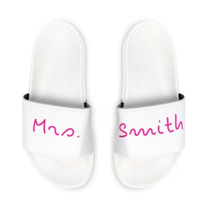 Op de afbeelding: Een paar witte slippers met roze tekst. De linker slipper zegt "Mrs." en de rechter slipper zegt "Smith."