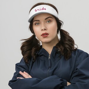 May include: A white visor with the word "Bride" written in pink script on the front. The visor is being worn by a person with brown hair styled in pigtails. The person is wearing a navy blue jacket.