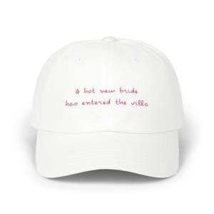 May include: White baseball cap with the text "A hot new bride has entered the villa" embroidered in pink.
