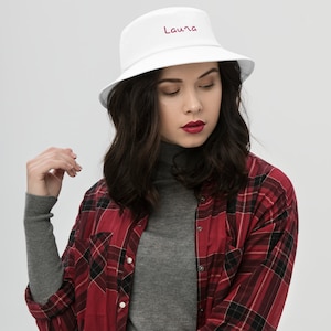 May include: White bucket hat with the name "Laura" embroidered in burgundy on the front. The hat is worn by a person with long brown hair and a red and black plaid shirt.