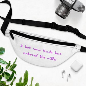 Könnte beinhalten: Eine weiße Bauchtasche mit einem schwarzen verstellbaren Riemen und einer violetten Aufschrift, die "A hot new bride has entered the villa" lautet.