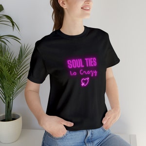 May include: A black t-shirt with a pink neon graphic that says "Soul Ties Is Crazy" with a pink heart and arrow.
