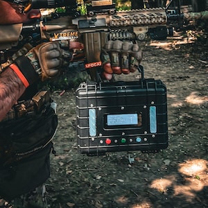 Pode incluir: Uma pessoa aponta uma espingarda com mira telescópica em uma área arborizada. A espingarda tem a palavra "XTREME". Uma caixa preta com tela digital e botões é segurada na frente da pessoa.