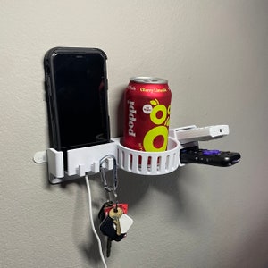 Puede incluir: Organizador blanco de pared con un soporte para teléfono, un portavasos, un gancho para llaves y un soporte para control remoto. El portavasos tiene una lata roja con el texto "Poppi" y "Cherry Limeade" en ella.