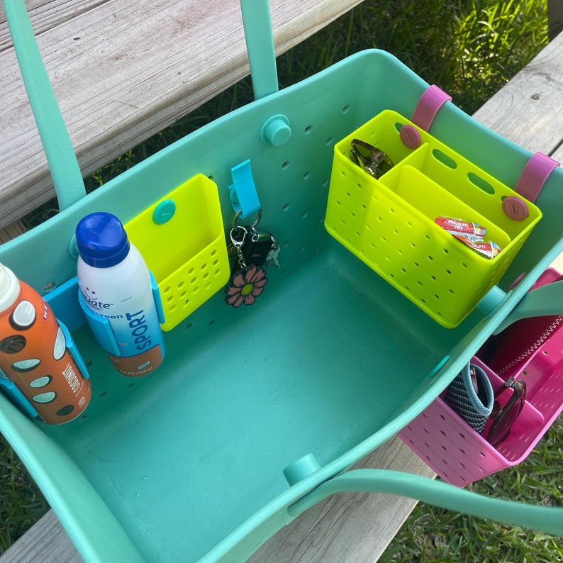 May include: A teal tote bag with multiple neon green and pink organizer inserts. The bag contains a bottle of sunscreen, keys, and other small items. The bag is sitting on a wooden surface.