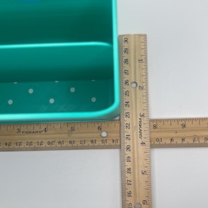 May include: A teal plastic organizer with a handle and two compartments. The organizer has white polka dots on the bottom. A wooden ruler is placed next to the organizer. The organizer is approximately 15 cm wide.