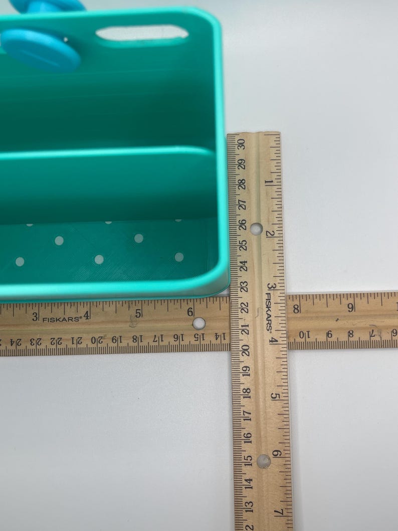 May include: A teal plastic organizer with a handle and two compartments. The organizer has white polka dots on the bottom. A wooden ruler is placed next to the organizer. The organizer is approximately 15 cm wide.