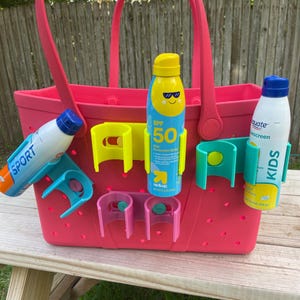 May include: A pink tote bag with attached holders for sunscreen bottles. The bag has a handle and is filled with sunscreen bottles. The sunscreen bottles are blue and white with yellow caps. The bag is sitting on a wooden surface.